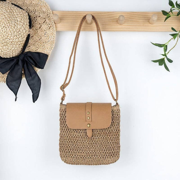 A Styled Collective straw crossbody bag with a tan faux leather flap and adjustable strap hanging on a wooden hook.