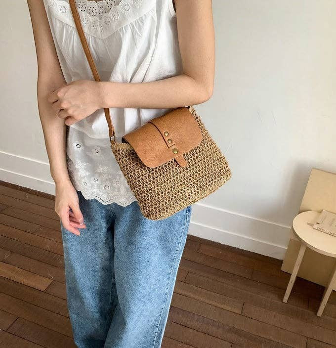A Styled Collective straw crossbody bag with a tan leather flap and gold button detail worn by a woman in jeans.